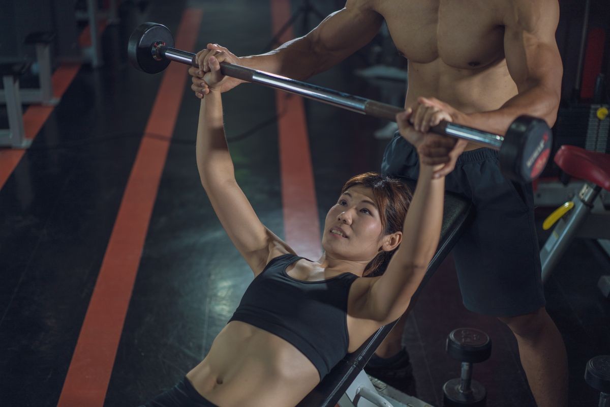 Personal trainer helping woman bench press in gym, Training with barbell, Personal trainer helping woman working with heavy dumbbells, Fitness instructor exercising with his client at the gym.